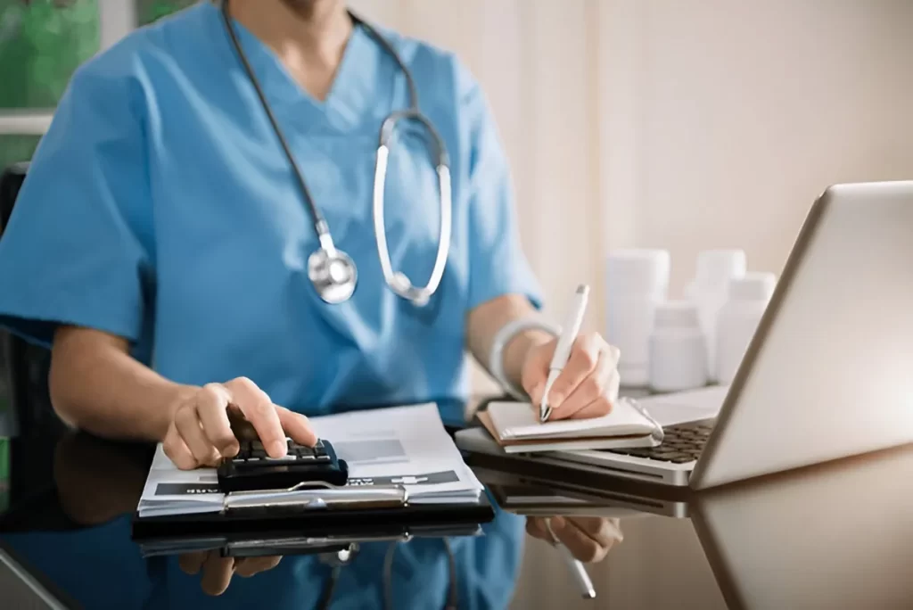 Nurse in scrubs using a calculator and taking notes beside a laptop, illustrating hospital billing services and medical expense management.